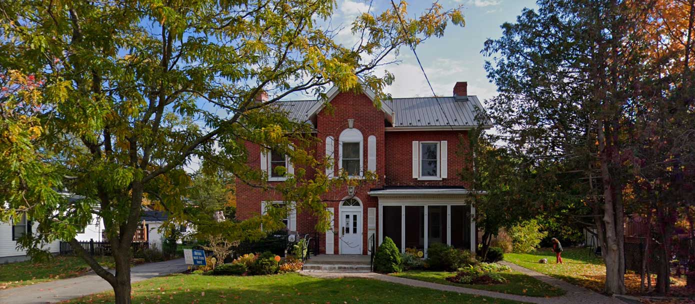 A two-story red brick house with a white front porch and a metal roof, surrounded by green and autumn trees.