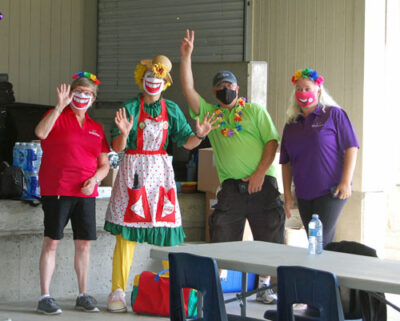 Four adults, including a clown, pose and wave while wearing oversized smiling mouth masks.