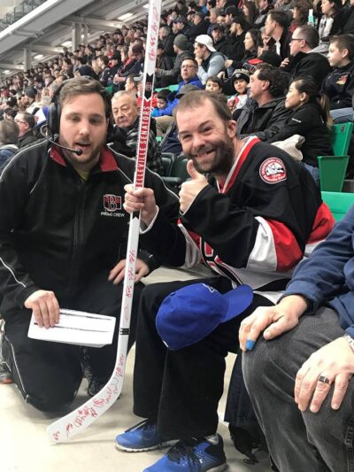 A person in a hockey jersey gives a thumbs-up while holding a signed hockey stick. Another person in a black promo crew jacket kneels next to them.