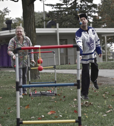 A man and woman play a game of ladder golf outdoors. The man, wearing a Toronto Maple Leafs jersey, is in the motion of throwing a bolo while the woman laughs in the background.