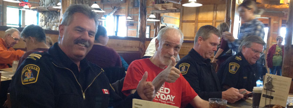 A person in a dark uniform with a Canadian flag on their shoulder smiles at the camera, sitting at a table in a restaurant. To their right, another person in a red t-shirt gives a thumbs-up. Two other people in uniforms are also visible at the table.